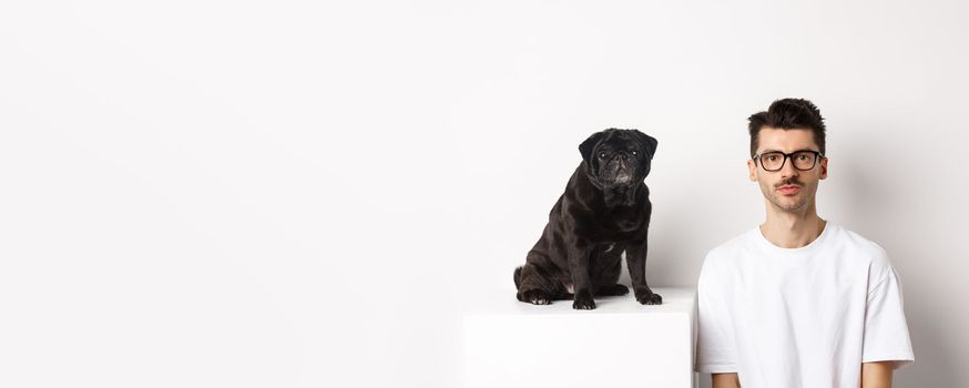 Image of handsome hipster in glasses sitting next to black cute pug dog, both staring at camera over white background.