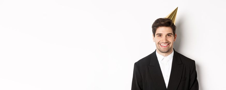 Close-up of handsome happy man in suit and party hat, smiling joyful, celebrating holiday, standing against white background.