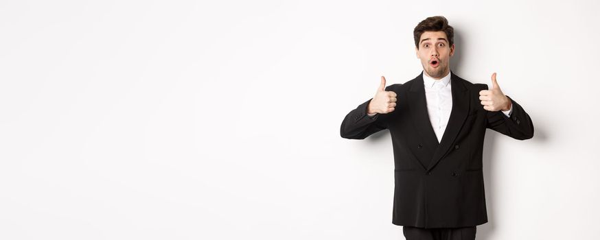 Concept of new year party, celebration and lifestyle. Portrait of excited handsome businessman in formal suit, showing thumbs-up in approval, recommending shop, standing over white background.