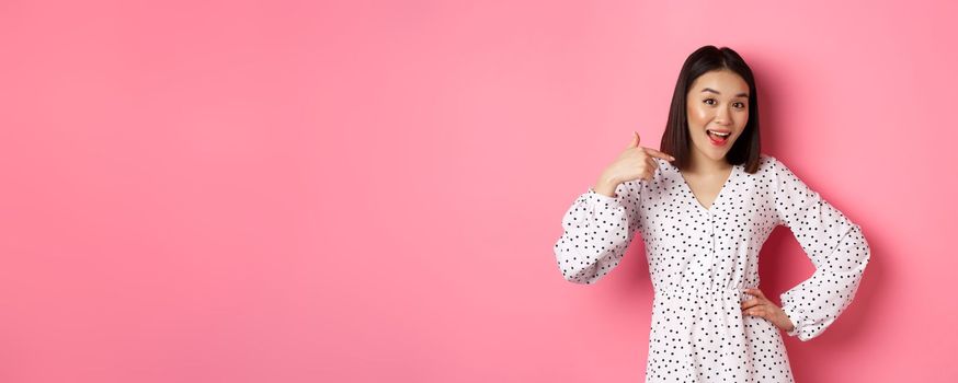 Beautiful asian girl in dress looking happy, pointing finger at herself, standing on romantic pink background.