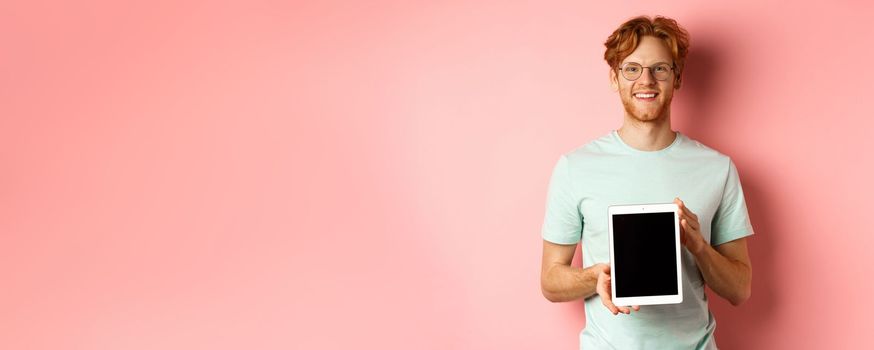 Funny redhead guy in glasses demonstrate digital tablet screen, smiling at camera, standing over pink background.