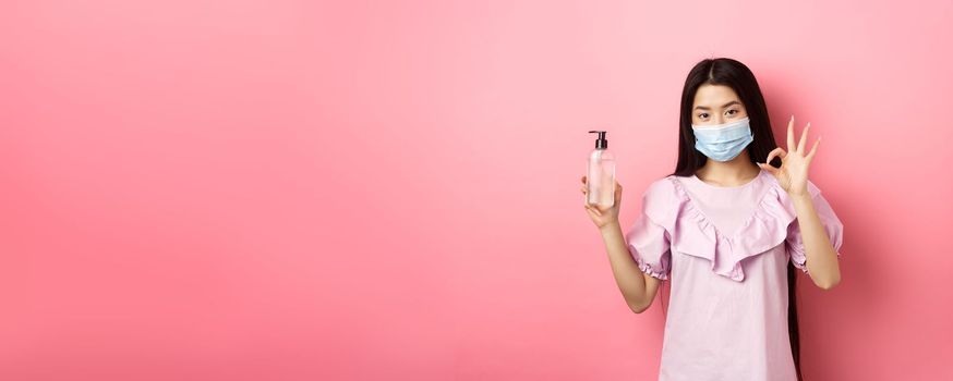 Healthy people and covid-19 pandemic concept. Cheerful asian woman in medical mask recommend hand sanitizer, showing okay sign, good antiseptic bottle, white background.