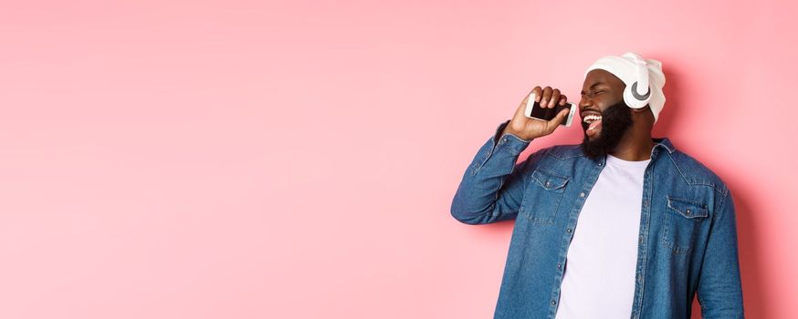 Carefree african-american man listening music in headphones, singing in mobile phone as microphone, standing over pink background.