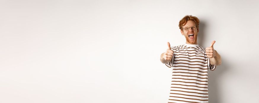 Happy redhead man scream yes and showing thumbs-up, approve and praise awesome company, standing over white background.