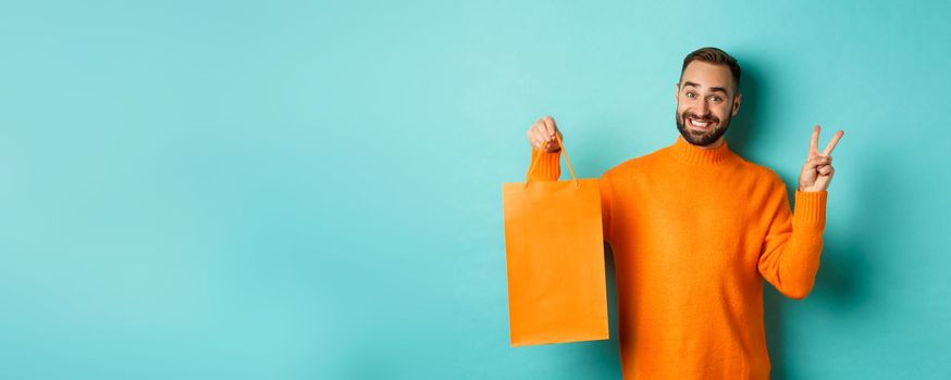 Happy young man showing peace sign and orange shopping bag, smiling pleased, standing over turquoise background. Copy space