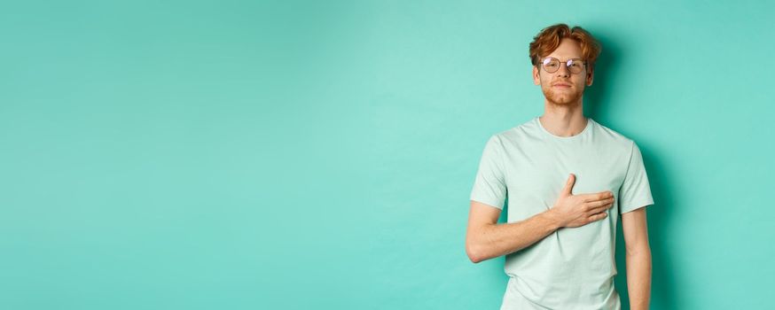 Proud young redhead man in glasses holding hand on heart, listening national anthem with respect, standing in t-shirt against turquoise background.