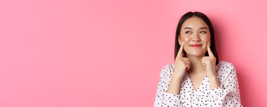 Beauty and skin care concept. Close-up of beautiful asian woman poking cheeks and smiling satisfied, standing over pink background.