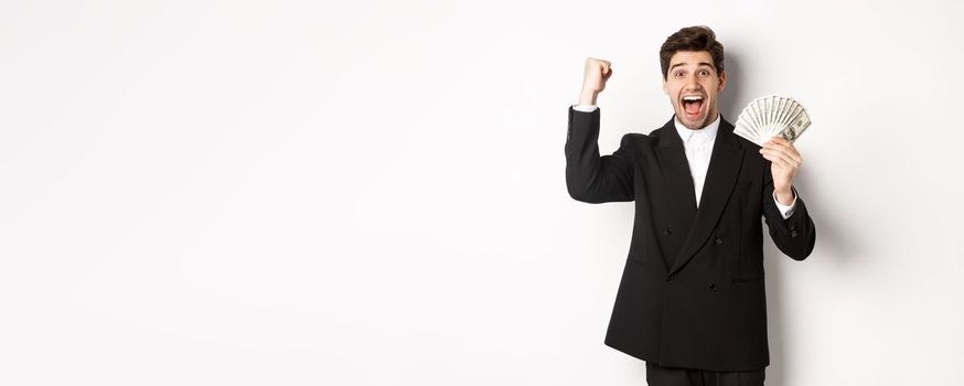 Portrait of handsome businessman in black suit, winning money and rejoicing, raising hand up with excitement, standing against white background.