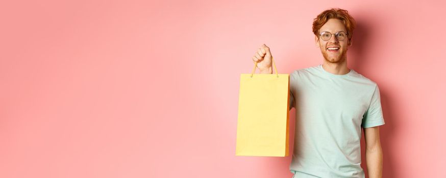 Handsome young man buying presents, holding shopping bag and smiling at camera, standing over pink background.