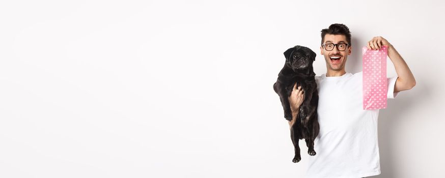 Image of hipster guy pet owner, holding cute black pug and dog poop bag, standing over white background.