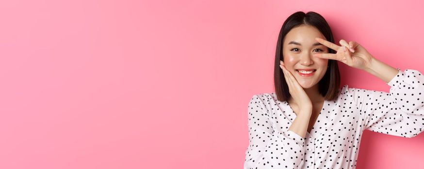 Beauty and lifestyle concept. Close-up of cute asian woman showing peace sign and touching cheek, smiling happy at camera, standing over pink background.