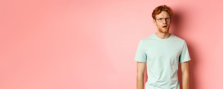 Portrait of handsome redhead man in glasses looking confused, open mouth and stare at camera, cant understand nothing, standing over pink background.