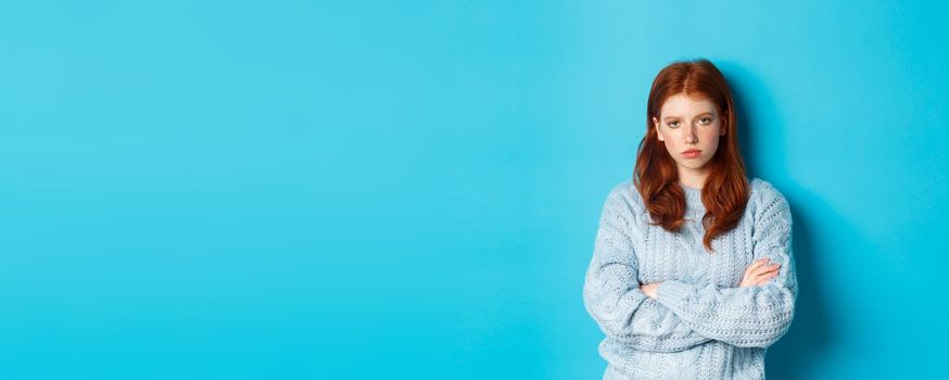 Annoyed and bothered redhead teeange girl cross arms on chest, staring at something lame and boring, standing against blue background.