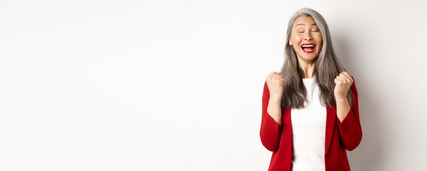 Relieved asian senior businesswoman making fist pump, saying yes and smiling satisfied, triumphing and winning, standing over white background.