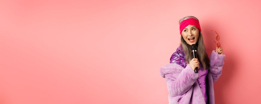 Gorgeous asian senior woman singing karaoke in mic, perform song and looking happy, standing over pink background.