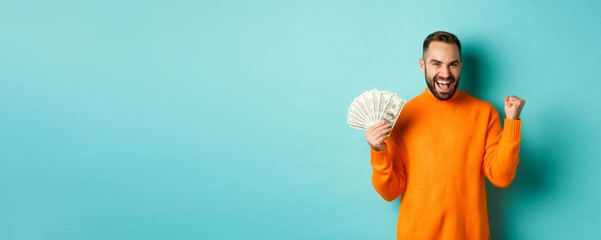 Shopping. Cheerful guy holding money, winning prize in cash and making fist pump, triumphing with satisfied expression, standing in orange sweater against turquoise background.