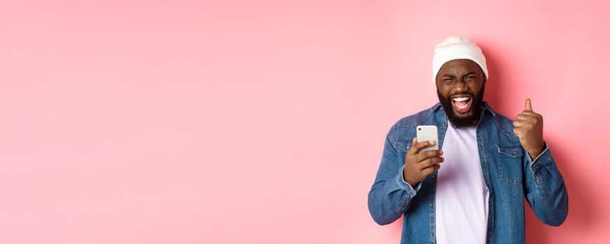 Technology and online shopping concept. Happy Black man rejoicing, winning in app, holding smartphone and scream yes, standing over pink background.