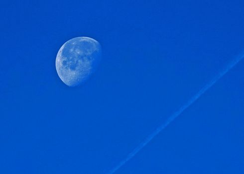 Waning gibbous moon with a jet stream in clear blue sky.