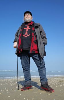 Senior man standing on the beach. He wears a shirt with a templars cross. It's beautiful sunny day in spring