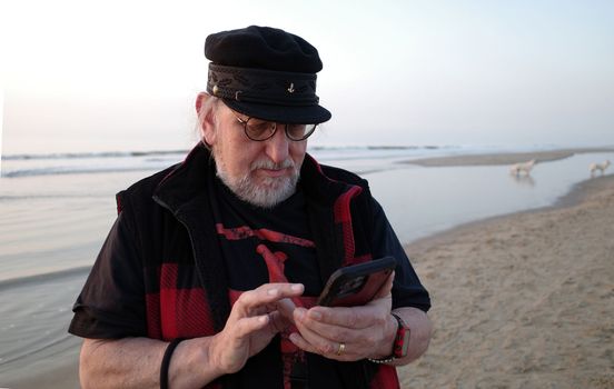 A senior man with captains cap, glasses and beard is busy with his smartphone. He's at the beach. Dogs are playing in the background