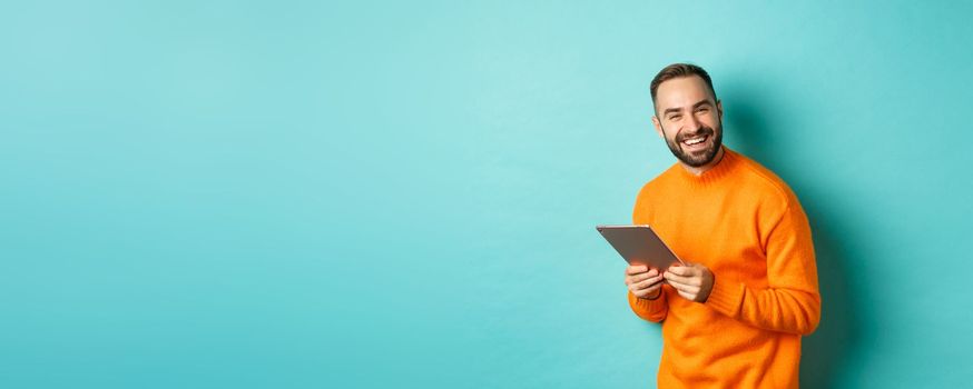 Handsome bearded man using digital tablet, laughing at camera, standing happy against light blue background.