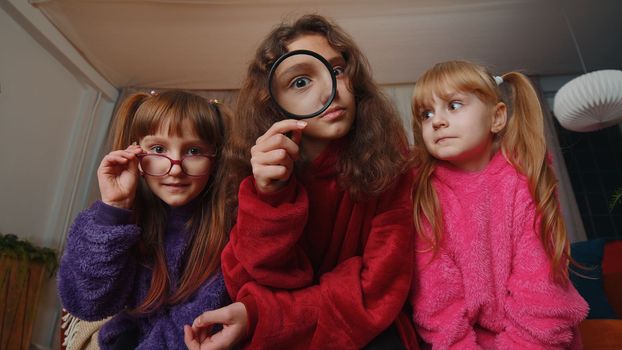 Investigator teenage girl and toddler child sisters holding magnifying glass near face, looking into camera with big zoomed funny eye, analysing, playing. Three siblings children kids at home playroom