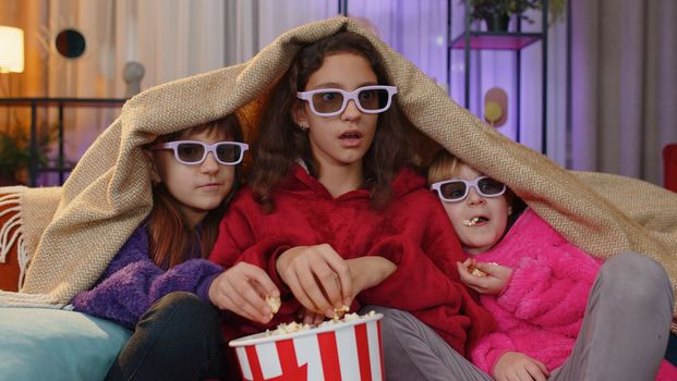 Excited teenage girl and little toddler child sisters under plaid eating popcorn and watching interesting tv serial, game, film, online social media movie. Three siblings children kids friends at home