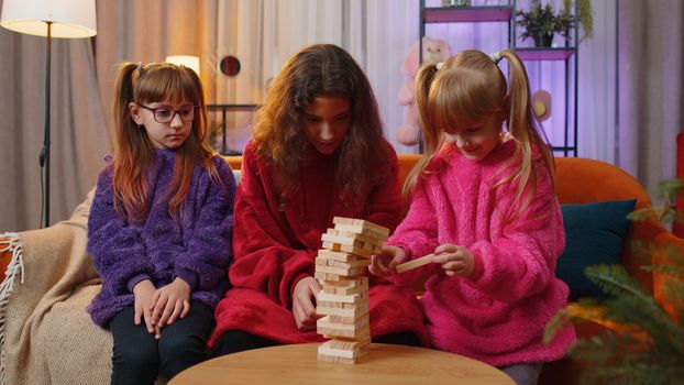 Teenage girl and toddler child sisters build tower from wooden bricks, losing board game competition. Three siblings children kids friends having fun with tower, playing with blocks at home playroom