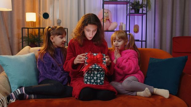 Happy teen girl and toddler child sisters opening Christmas gift box with excited surprised face. Female three children kids siblings or friends holding light glowing birthday present at home playroom