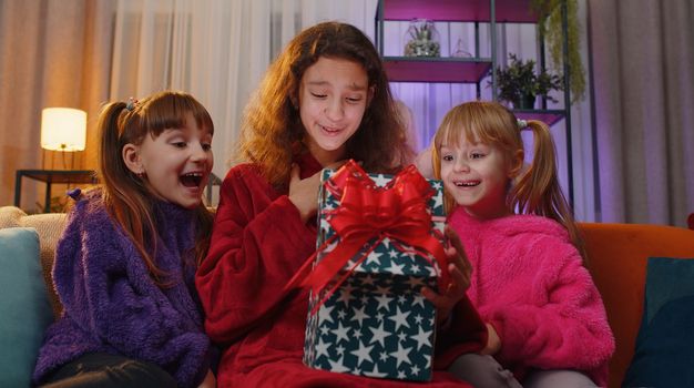 Happy teen girl and toddler child sisters opening Christmas gift box with excited surprised face. Female three children kids siblings or friends holding light glowing birthday present at home playroom