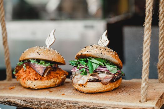 Delicious burgers on a wooden base. Two delicious homemade burgers of beef, cheese and vegetables on an old wooden table. Fat unhealthy food close-up. Burger fest.