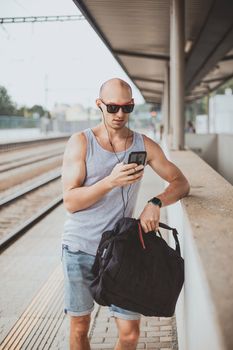 Young man in a t-shirt on the platform waiting for a train using mobile phone. Man by train station platform suburban with bag and mobile phone. Traveling by train