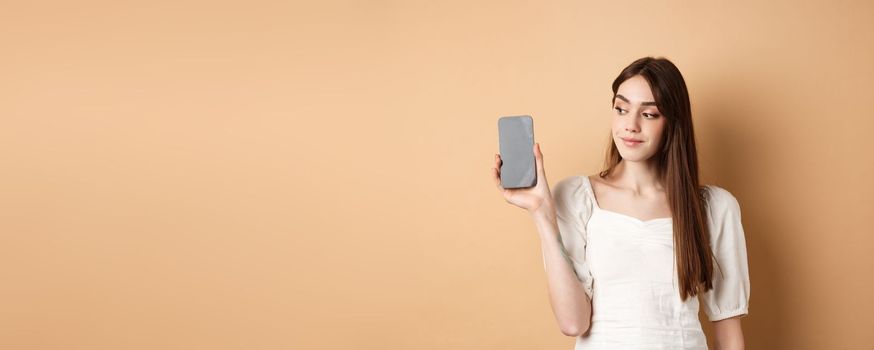 Attractive young woman showing empty smartphone screen, smiling and looking aside, standing on beige background.