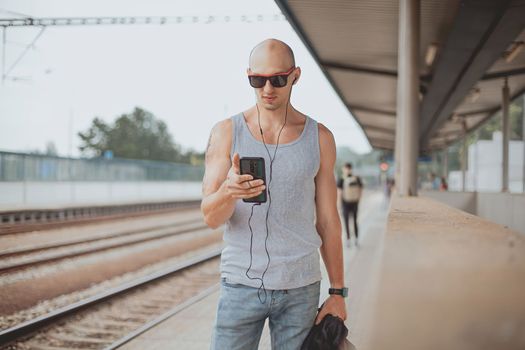 Young man in a t-shirt on the platform waiting for a train using mobile phone. Man by train station platform suburban with bag and mobile phone. Traveling by train