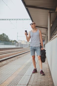 Young man in a t-shirt on the platform waiting for a train using mobile phone. Man by train station platform suburban with bag and mobile phone. Traveling by train