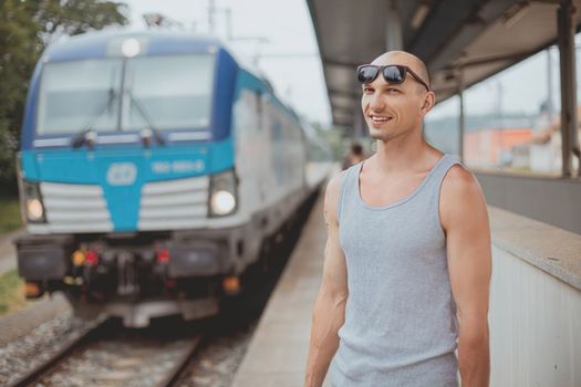 Young man in a t-shirt on the platform waiting for a train using mobile phone. Man by train station platform suburban with bag and mobile phone. Traveling by train