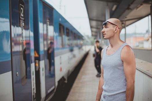 Young man in a t-shirt on the platform waiting for a train using mobile phone. Man by train station platform suburban with bag and mobile phone. Traveling by train