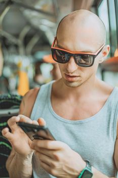 Young mansmartphone travelling by bus in city. A young man wearing a T-shirt and sunglasses uses a mobile phone on public transport. A man on a bus with a mobile phone.