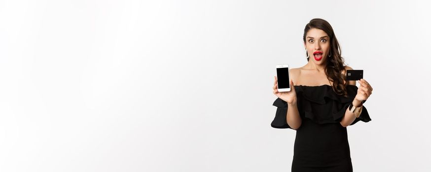 Fashion and shopping concept. Amazed beautiful woman showing smartphone screen and credit card, looking excited, buying online, white background.