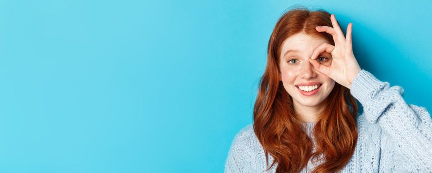 Headshot of cute teenage redhead girl showing okay sign on eye and smiling, standing happy against blue background.