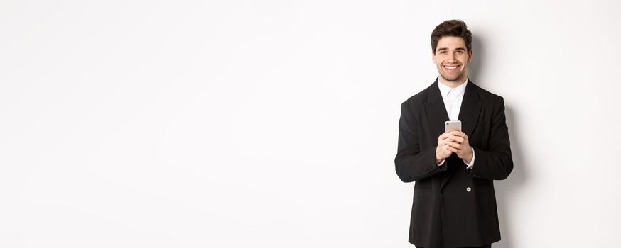 Portrait of successful, handsome businessman in suit, writing message on smartphone and smiling, standing over white background.