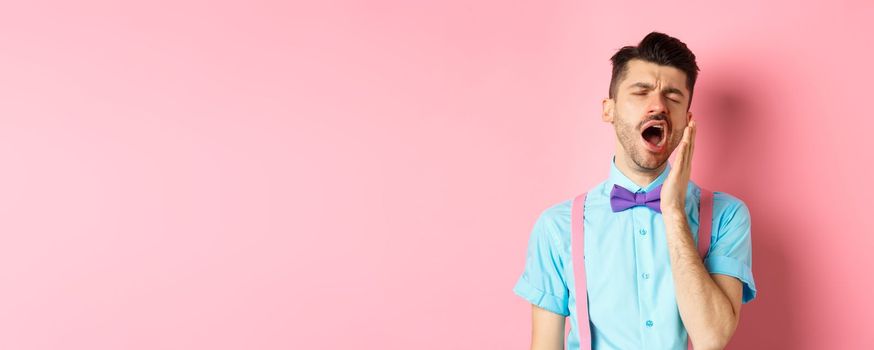 Image of tired guy in bow-tie yawning with opened mouth and closed eyes, standing sleepy, feeling fatigue, standing over pink background.