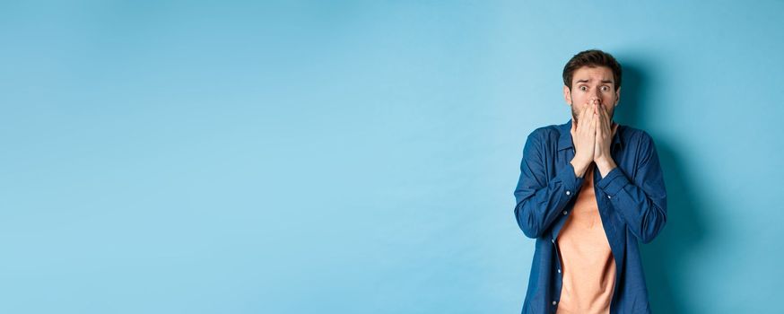 Scared anxious man covering mouth with hands and looking at something terrifying, standing on blue background.