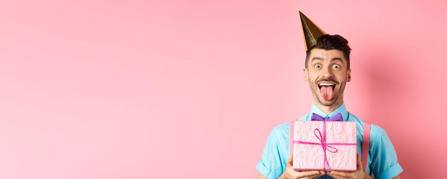 Holidays and celebration concept. Funny guy celebrating birthday, wearing party hat, holding b-day gift and showing tongue with happy face, pink background.
