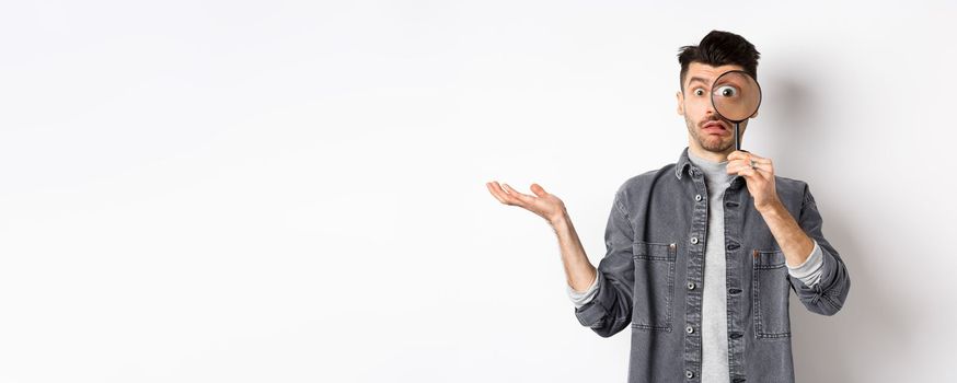 Confused man look through magnifying glass and shrugging clueless, standing on white background.