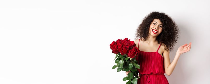 Happy woman celebrating, wearing stylish dress and holding flowers, smiling at camera, white background.