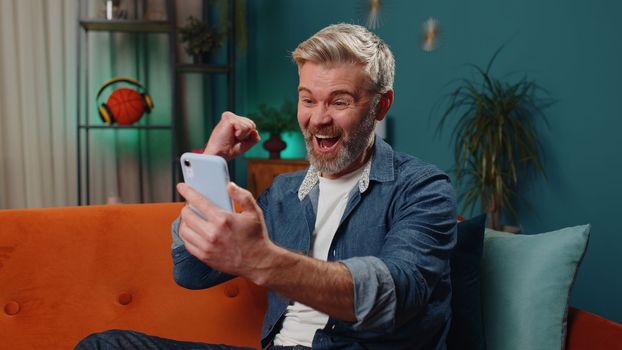 Middle-aged old man use mobile smartphone typing browsing say Wow yes found out great big win good news celebrate lottery jackpot doing winner gesture. Mature guy at home in night room sitting on sofa