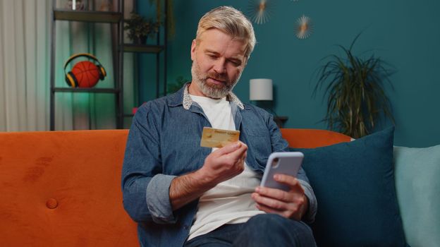 Middle-aged old man using credit bank card and smartphone while transferring money, purchases online shopping, order food delivery at home apartment indoors. Mature senior guy in evening room on couch