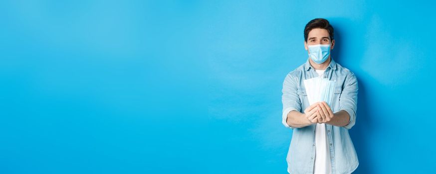 Concept of coronavirus, quarantine and social distancing. Young man giving medical mask to prevent virus spread, standing against blue background.