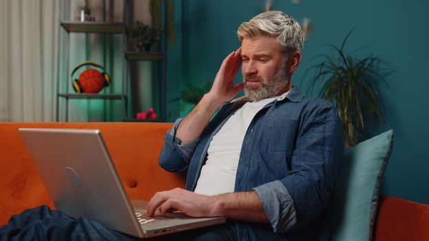 Tired middle-aged man freelancer use laptop, suffering from headache problem tension and migraine, stress at home. Mature guy works on notebook, sends messages, makes online purchases, watching movies